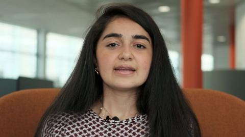 A young woman looking at the camera, she has long dark hair and a patterned black and white top.