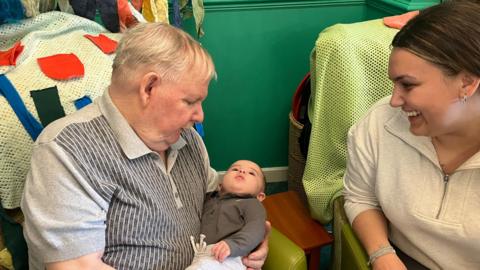 Pat, an older man, on the left, cradling baby Huxley, while Huxley's mother smiles and watches them both