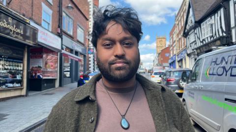 Sho Abdul is stood on Whitchurch high street and is looking into the camera. He is wearing a light brown T-shirt with an olive green wool jacket over the top, and is wearing a necklace with a large blue gemstone pendant. He has short dark hair and a short beard. Abdul is framed by shops along the edges of the photograph, with a church tower visible in the background. The sky is blue with some white clouds. 