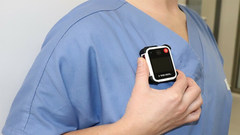 Close up generic image of a body-worn camera on a female nurse in blue nurses uniform. Her hand is clutching the device.