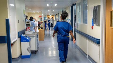 A nurse wears blue scrubs as she walks down a hospital corridor, there are other nurses in the background doing different tasks.