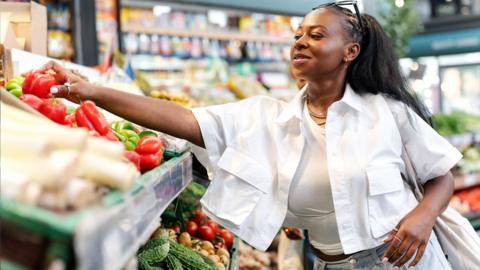 A woman reaches for a pepper on a shelf in a supermarket