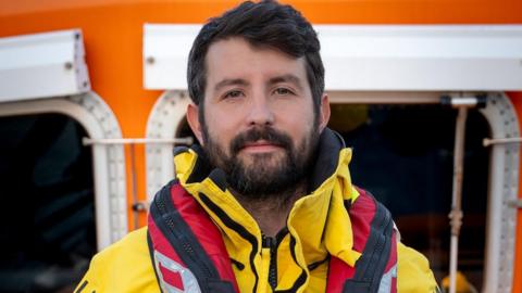 Will Roberts is smiling at the camera, wearing his RNLI lifeboat crew uniform which is a yellow anorak and a red lifebelt. He has black hair, a black beard and brown eyes. Behind him is the lifeboat wheelhouse which is orange with white trim.