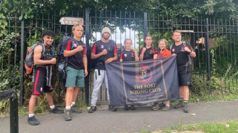 three boys and two girls are standing in front of a black iron fence, next to a woman and a man. They are holding a black flag that reads "the fort boxing club". they have their hands raised with their fists clenched 