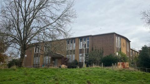 A building at the top of a mildly sloped grass bank. The brick building is large with windows. There are a number of windows.