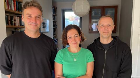 Two men and a woman stood in a living room looking directly at the camera