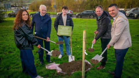 John's family members are pictured with mayor Ruairí McHugh holding shovels near the sapling. (L_R) Erin Gallagher, Jim Gallagher, Tiarnán Gallagher, John Donaghy and mayor Ruairí McHugh)
Mayor Ruairí McHugh) is wearing his mayoral chain in a cream suit jacket and navy trousers. Erin Gallagher (pictured far left) is in a dark leather jacket and navy trousers. She has long curly red hair. Jim Gallagher is in a navy coat and holding a plaque near the sapling. 