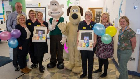 Wallace and Gromit characters wave at the camera on a ward at the Bristol Royal Children's Hospital. They are surrounded by members of staff, some of which are holding balloons and boxes of cupcakes.