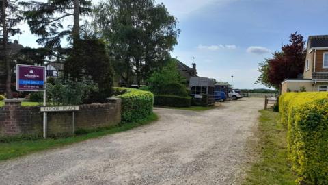 A gravel path with hedges and houses on either side. There is a 'For Sale' sign outside the nearest home above a road sign which reads 'Tudor Place'. There are also several trees and a horse box visible. 