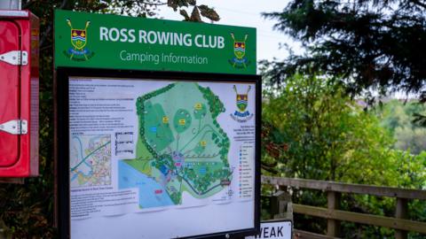 A large green information board for Ross Rowing Club stands beside a wooden footbridge, surrounded by trees and greenery.