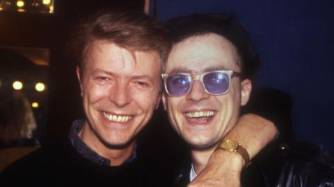 David Bowie and guitarist Kevin Armstrong. David is on the left, Kevin is on the right. Kevin is white and wearing blue-tinted glasses. Both are smiling.