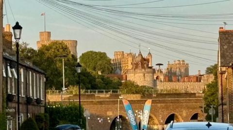 Windsor Castle is bathed in the golden setting sun of golden hour. It sits above a row of terraced houses and the union flag flies high above the castle on a flag pole. The sky is clear and blue. Telephone lines to houses cross through the shot.