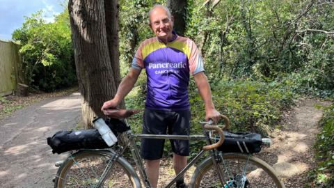 Steve Groves wearing a purple Pancreatic Cancer UK cycling top holding his chrome coloured 90 year old bicycle standing in a woodland area