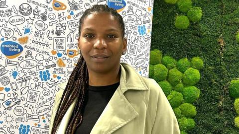 A medium close up picture of a lady called Luana wearing a black top and green jacket. She is stood in front of a green, garden wall and a promotional poster board for Enabled Living. 