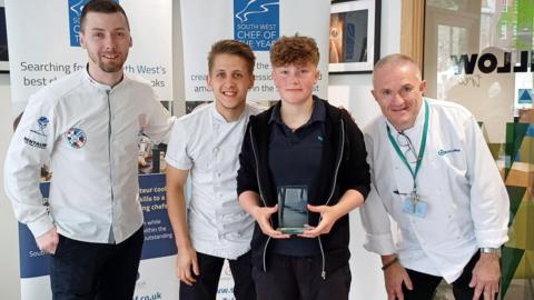 Three judges wearing white chef's coats standing with Lexie Colley in the middle. Lexie is wearing a navy blue polo and black jacket and she's holding the trophy in her hand.