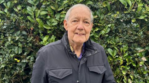 Paul is standing in front of a green bush. He is wearing a charcoal coloured jacket. He is smiling slightly.