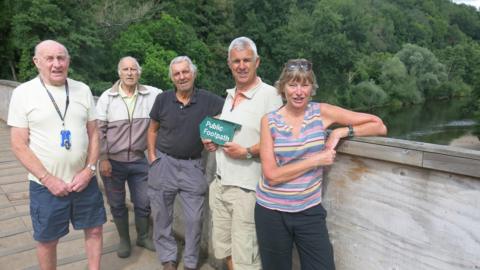 Image of councillors standing to the side of a bridge over some water. They are learning on the wooden side, wearing shots and t-shirts in seemingly warm weather. 