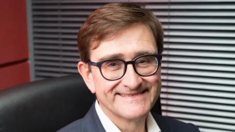 A man with glasses and brown hair sat in an office chair smiling into the camera.