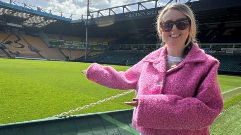 A woman with blonde hair can be seen wearing a pink jacket and sunglasses as she points to a football pitch behind her and the stands alongside it.