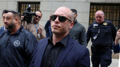 Gannon Van Dyke, with a shaved head and wearing sunglasses, as well as a blue suit jacket and black shirt, arrives at court surrounded by police and media. 