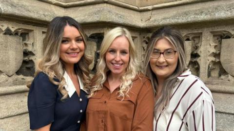 Louise Hubbard, Justine Ducker and Danielle Mann smiling at the camera. Louise is wearing a blue dress with white buttons and a white collar. Justine is wearing a brown shirt. Danielle is wearing a red and white striped shirt and wears glasses