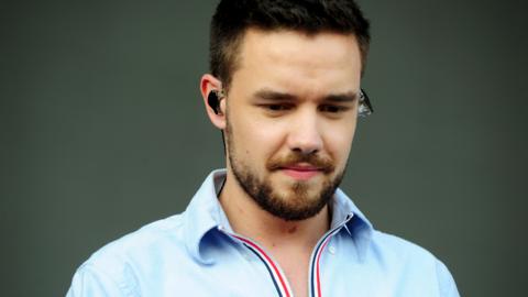 Liam Payne looks down onstage with earbuds in and wearing a blue zip up shirt/coat, in Chicago in 2018
