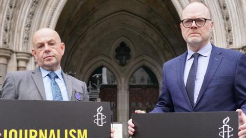 Journalists, Barry McCafferty on the left holding a sign that says 'Journalism is not a crime', wearing a grey suit with a light blue shirt and dark blue tie. Trevor Birney is standing on the right holding the same sign, wearing a navy suit, with a lilac shirt and purple tie