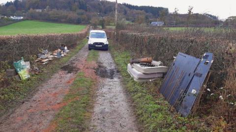 A country lane is pictured with mattresses, building waste and metal sheets piled on the grass verges on either side. A white van is parked on the road in shot.