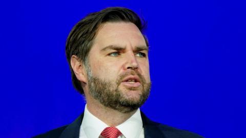 JD Vance wears a dark suit and red tie with a blue background behind him