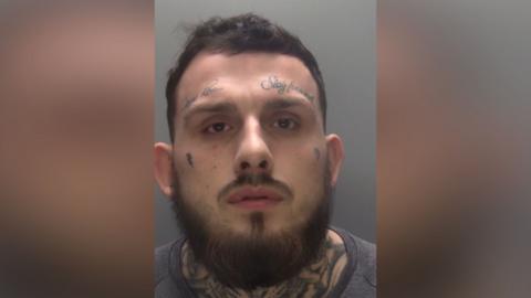 A man with tattoos above his eyebrows and neck looks at the camera in his police custody photo. He has a thick beard. He is wearing a dark T-shirt.