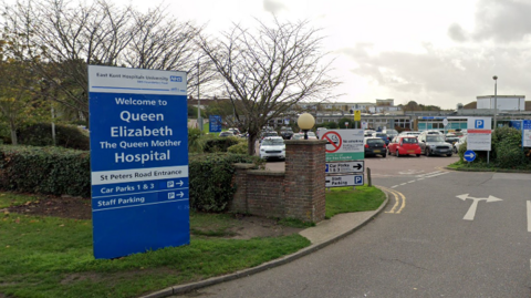 The main road entrance to the Queen Elizabeth the Queen Mother car park, with a blue sign to the left of the hospital's name