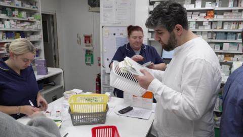 The image shows the inside of a pharmacy or dispensary area. There are several people working at a counter, which is covered with prescription bags, paperwork, and baskets in different colours (yellow, white, and red). One person is holding a white basket filled with medication boxes and checking the contents. Behind the counter, there are shelves stacked with various medicines and pharmaceutical products. A mobile phone and some documents are also visible on the counter. 