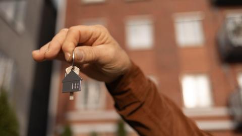 A hand holds up a key and a keyring in the shape of a house. A block of flats is in the background.