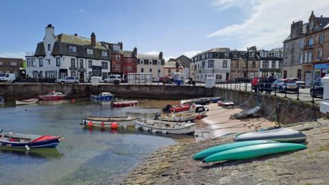 Millport on the Isle of Cumbrae. There are small boats moored in the harbour and colourful buildings along the main street. Cars are parked along the street and pedestrians walk along the pavement. 