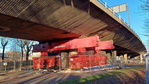 A large red metal structure props up the grey concrete flyover road. The equipment is surrounded by grey fencing.