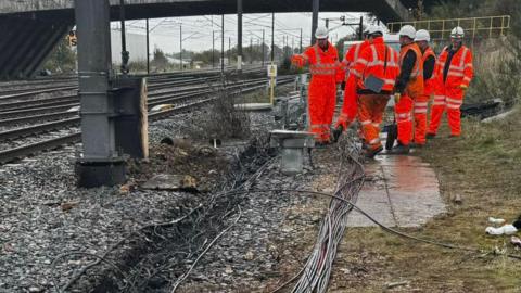 Engineers, in orange high vis clothing by a side of a railway track with damaged cables