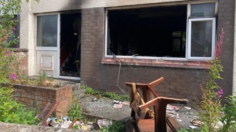 A house after a fire. The window has been smashed and door is gone. A room and the stairs are burnt. Some chairs and debris is lying outside i the garden.  