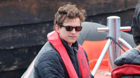 A close up of Tom Holland, wearing sunglasses and a red life jacket over a dark jacket, is sitting in an inflatable boat. He has his brown hair in his trademark tousled style. Behind him is a grey harbour wall. 