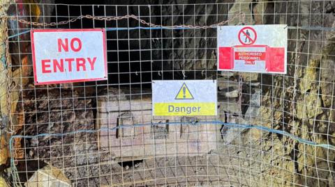 A metal fence blocks off a mine shaft. The fence has three signs on it - one says no entry in red letters on a white background. One says danger in black letters on a yellow background below a yellow triangle 'danger' symbol. The final sign says authorised personnel only. On either side of the mine shaft are rock walls. The shaft continues into darkness in the background.