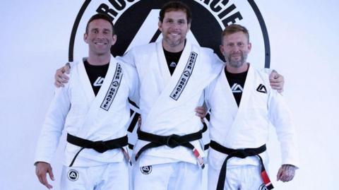 Three men wearing white gis standing shoulder to shoulder, in front of a club logo. All three have black belts.