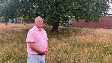An older man  wearing a pink short sleeved shirt, light coloured trousers and glasses stands with his hands clasped in front of his stomach. He is stood on an overgrown green space in a residential area, which is brown and full of weeds and tall grass. There is a large cherry tree and a house behind him.