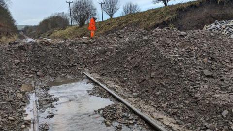 An engineer in orange hi-vis standing on top of piles of debris on top of a flooded railway track.