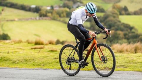 A man cycling up the hill with a background of fields and greenery behind him