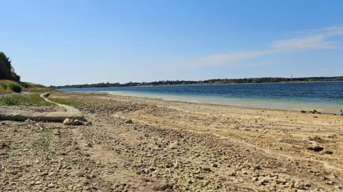 A view of Grafham Water lake and reservoir from the perspective of someone standing on the shore. It shows low water levels as the shore is widely exposed with many stones on . The water is turquoise then a deep blue, and there are pylons in the distance. To the left is a grassy bank and trees. 