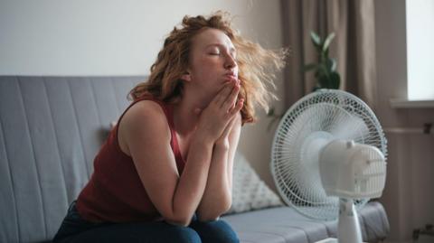 A young woman with fair skin and strawberry blonde hair is sitting bent forward on a sofa with her elbows on her knees and her hands beneath her chin. In front of her face is a fan. Her eyes are closed and her hair is blowing in the breeze of the fan. 