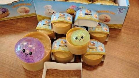 Piles of squishy toys on a table. They are sparkly and in the shape of bao buns. They have smiley faces on them and each one is contained in a little round box.