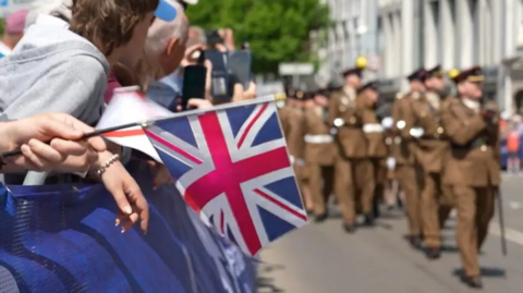The image shows spectators lining a street behind barriers, with one person holding a Union Jack flag in the foreground, while a group of uniformed service personnel march past in formation during a parade.
