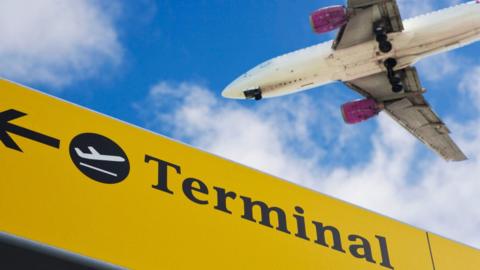 Plane flying over Stansted airport, with a sign to the terminal in the foreground.