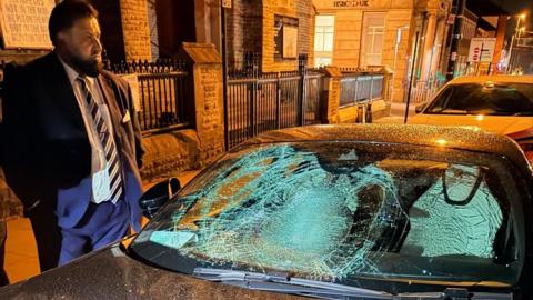 A man in a suit and tie with a beard and short hair stands next to a car parked on the roadside. The black car has a badly smashed up windscreen.