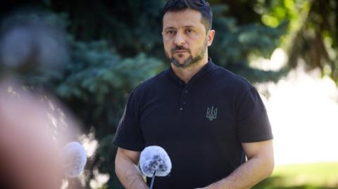 Zelensky, wearing a black polo shirt adorned with the Ukrainian trident looks on as he is photographed in a front of a microphone delivering a press conference outside 